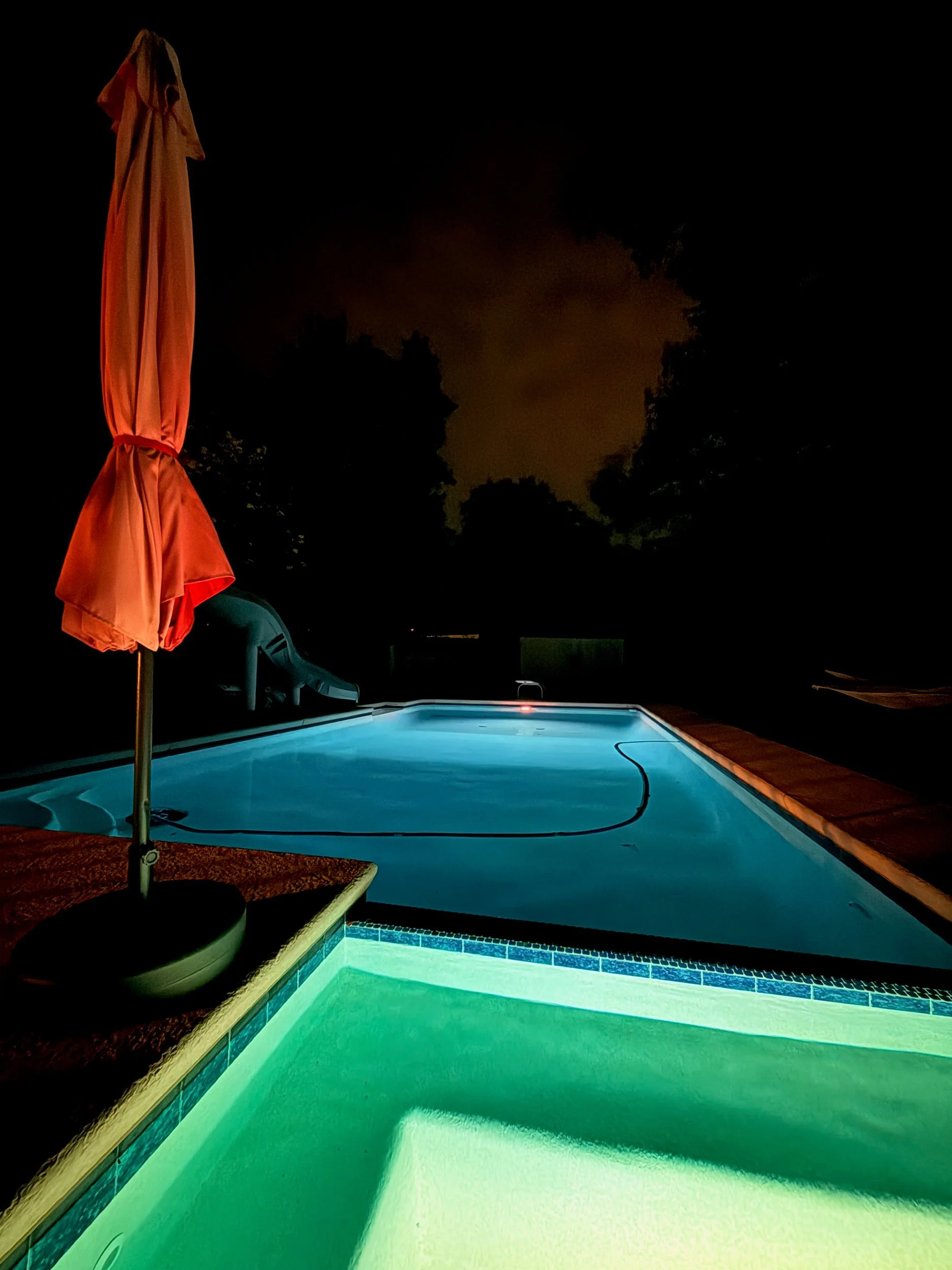 Swimming pool glowing turquoise at night beside a closed red umbrella