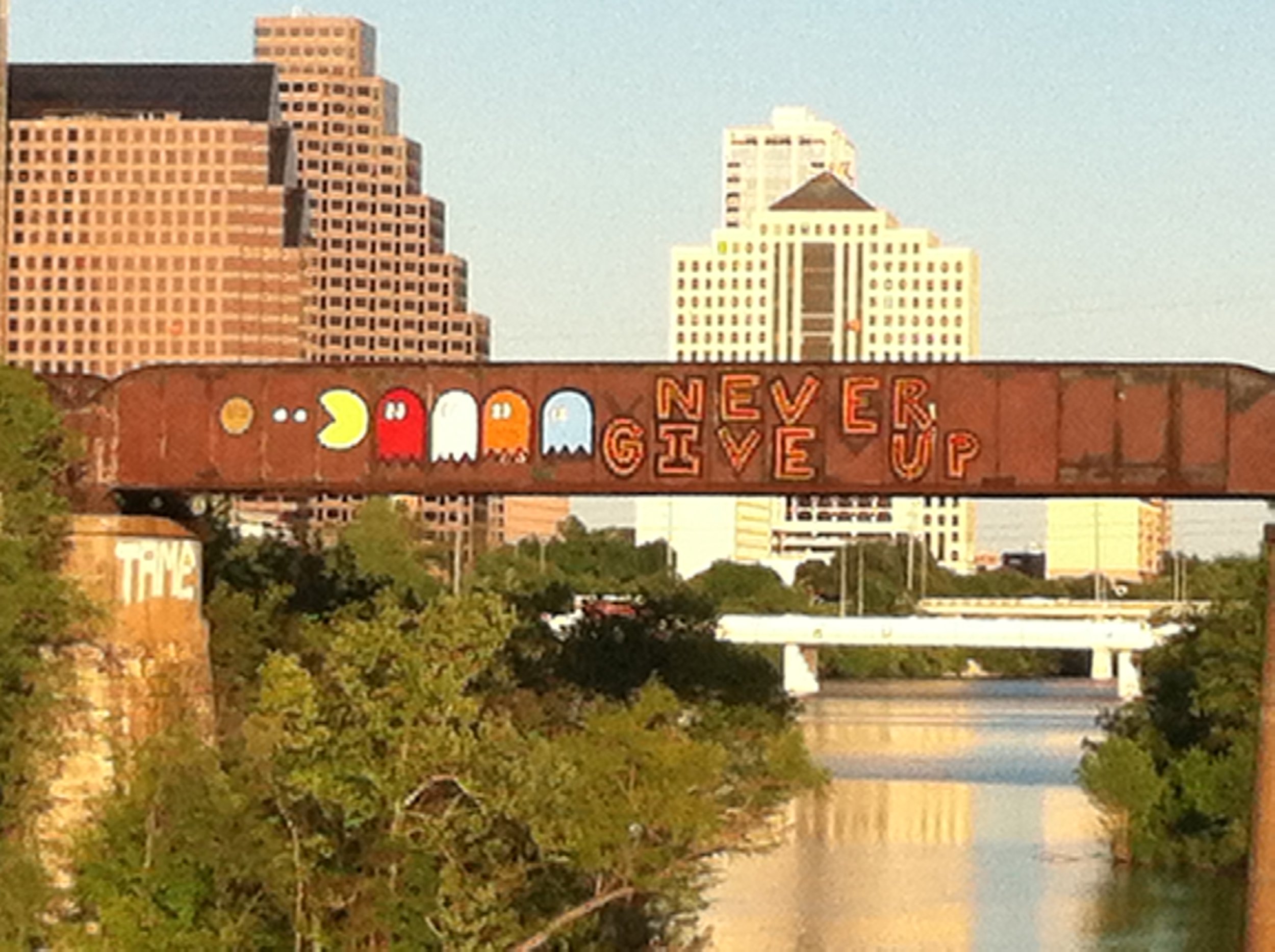 A billboard with colorful Pac-Man characters and the words "Never Give Up" in red letters, with a city skyline of tall buildings in the background.