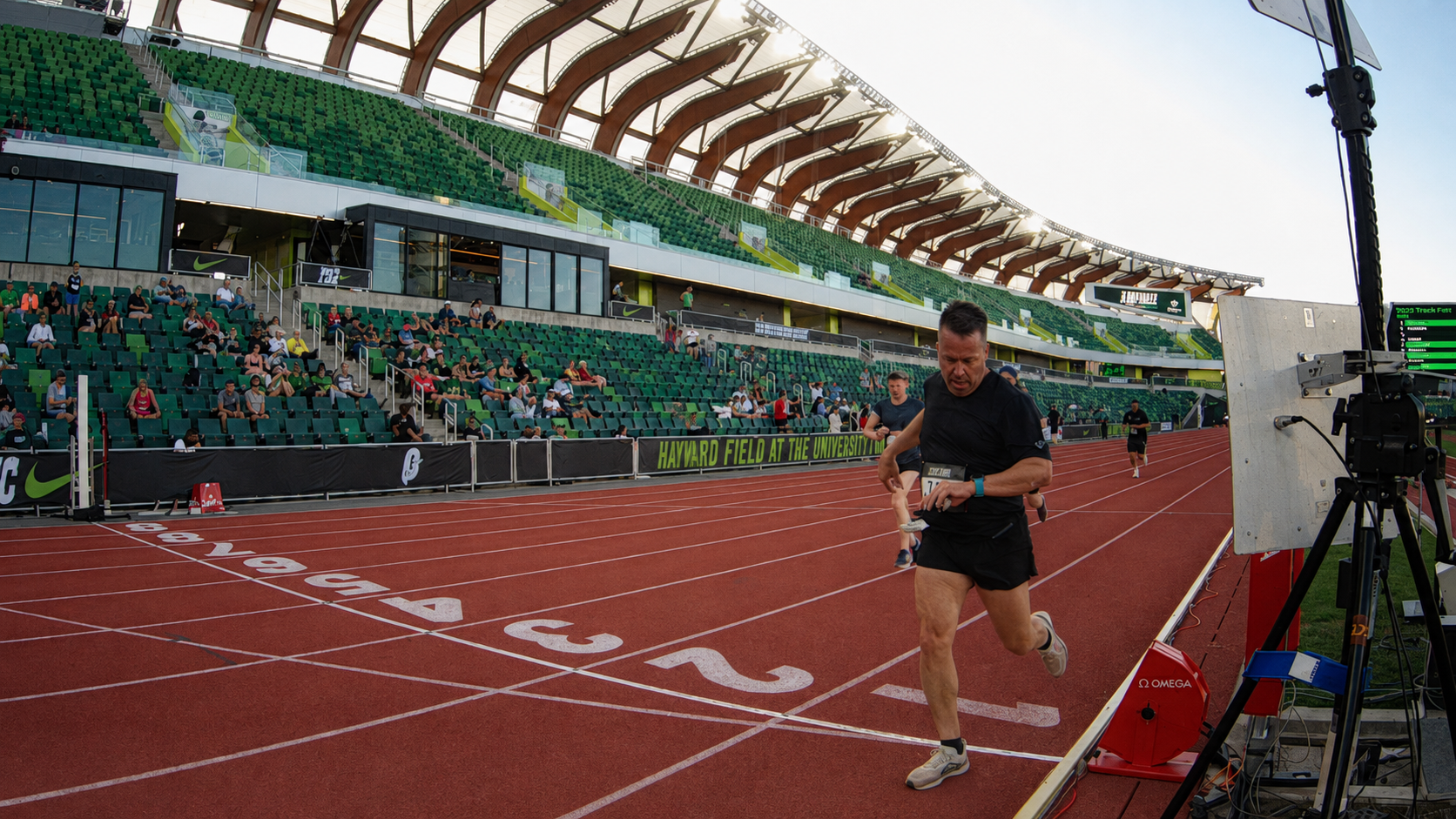 A male runner in black athletic clothing crossing the finish line at an outdoor track stadium during a race, with spectators seated in the stands and equipment on the side.