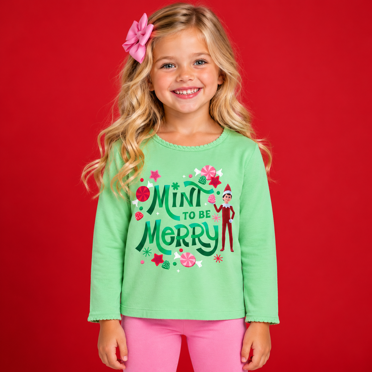 Smiling young blonde girl wearing a mint green long-sleeve Christmas shirt featuring a colorful “Mint to Be Merry” graphic with a cheerful Scout Elf, peppermint candies, and festive hand lettering, paired with pink leggings and a matching pink hair b