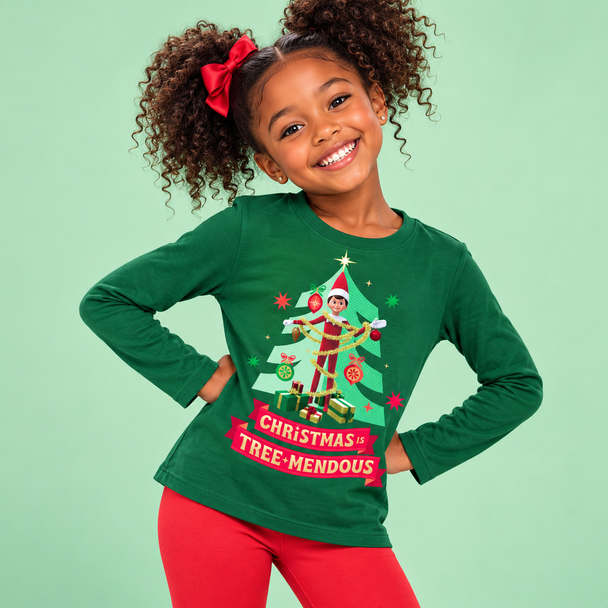 Smiling young girl wearing a green long-sleeve Christmas shirt featuring a festive elf, decorated Christmas tree, and "Christmas Is Tree-Mendous" graphic, paired with bright red leggings.