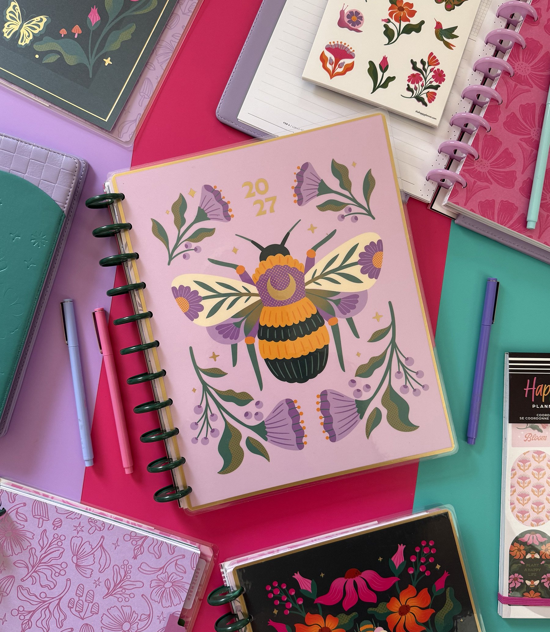 Colorful planners with floral and butterfly designs, placed on a purple, pink, and teal surface. Some planners have tabs and spiral bindings, with pens nearby.