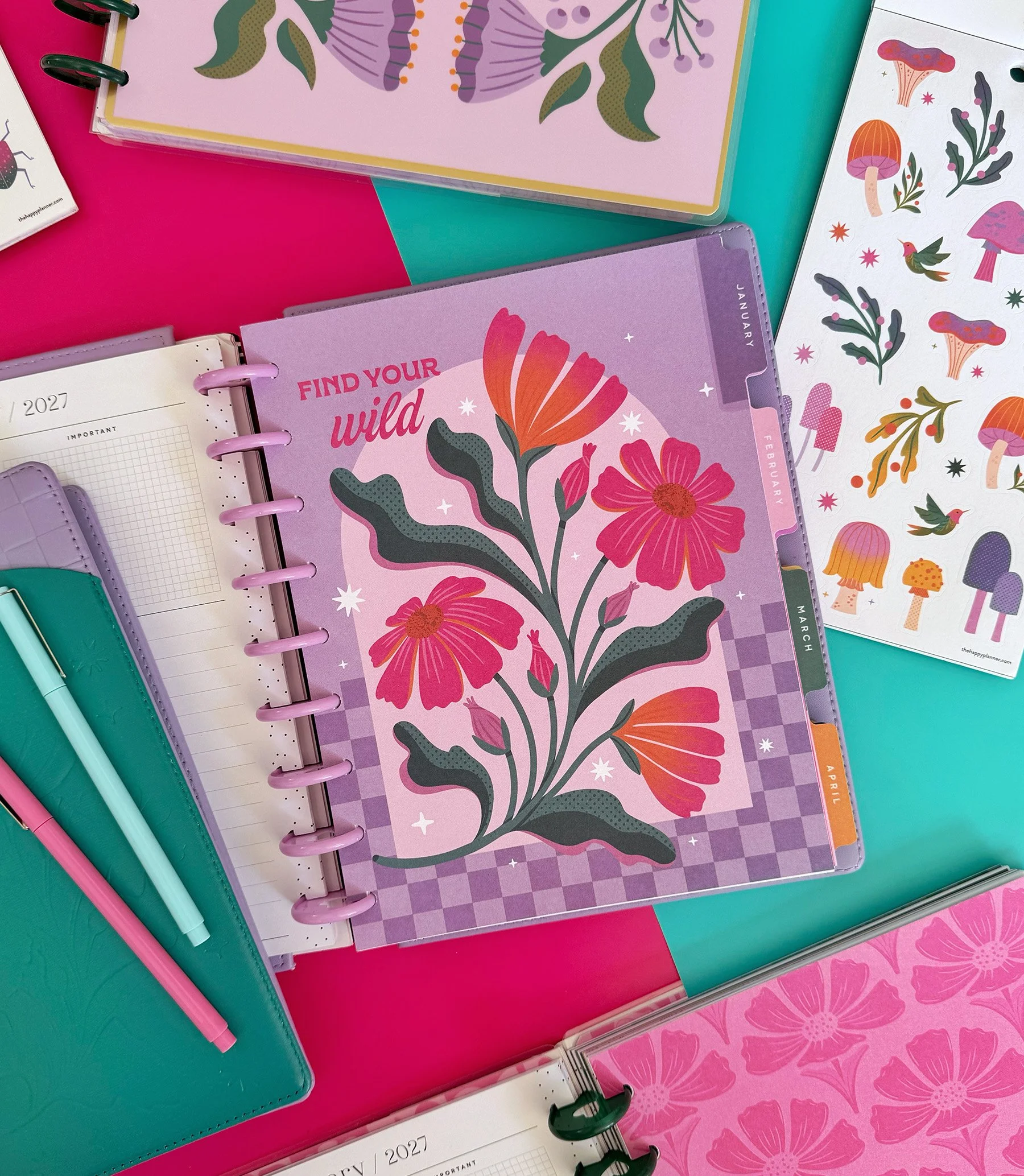 Colorful planners with floral and butterfly designs, placed on a purple and teal surface. Some planners have tabs and spiral bindings, with pens nearby.