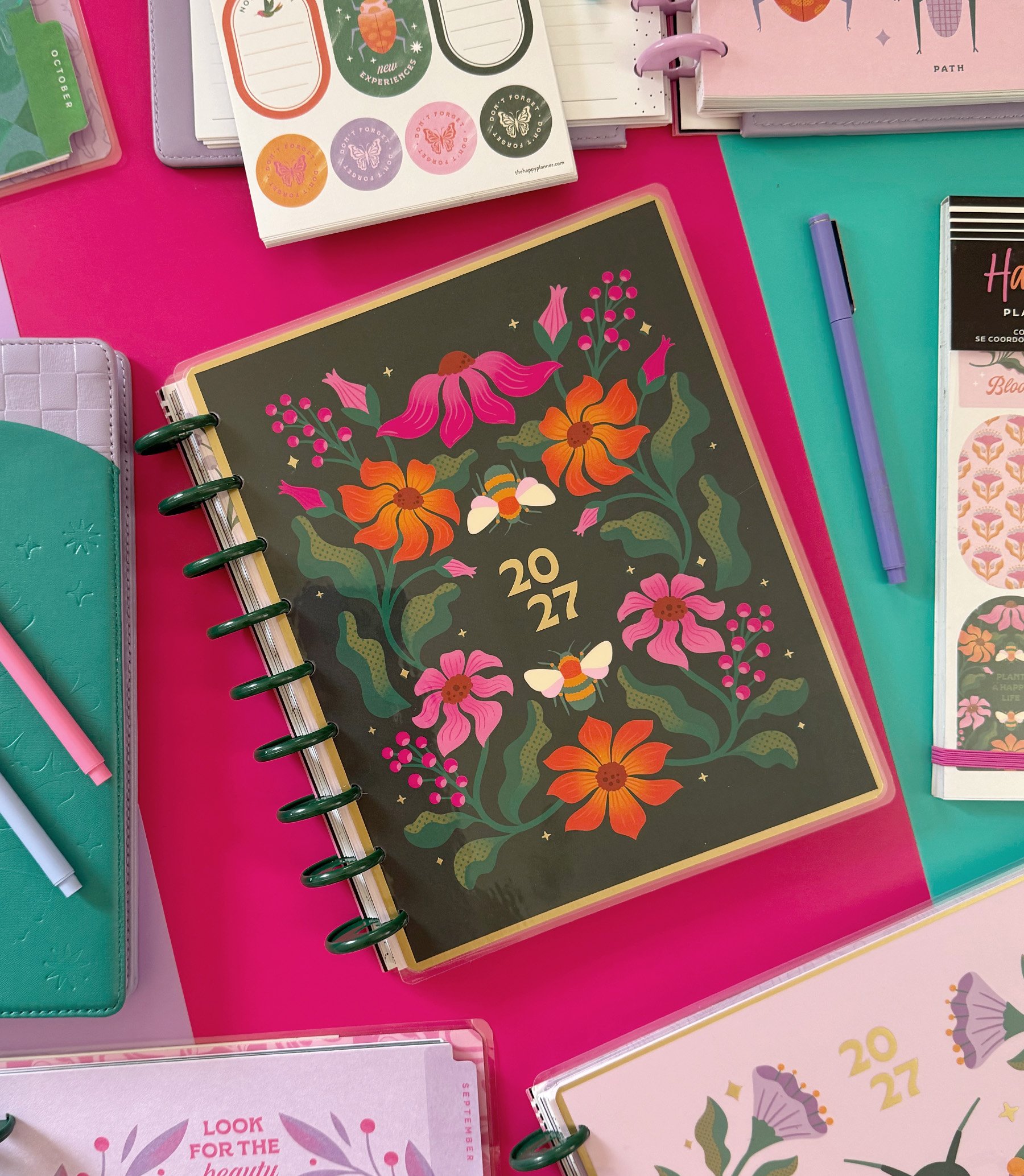 Colorful planners with floral and butterfly designs, placed on a pink and teal surface. Some planners have tabs and spiral bindings, with pens nearby.