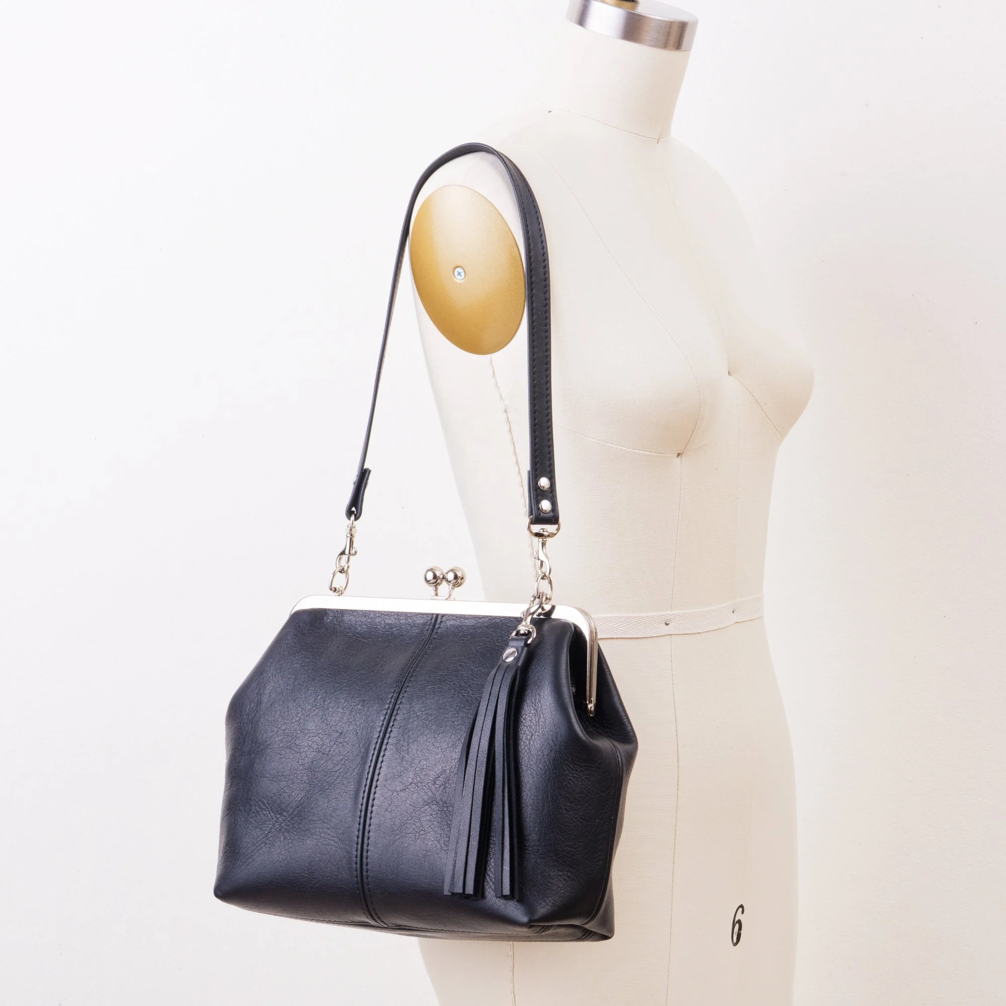 Black leather kiss lock handbag with silver finish hardware, standard shoulder strap and tassel charm on mannequin.