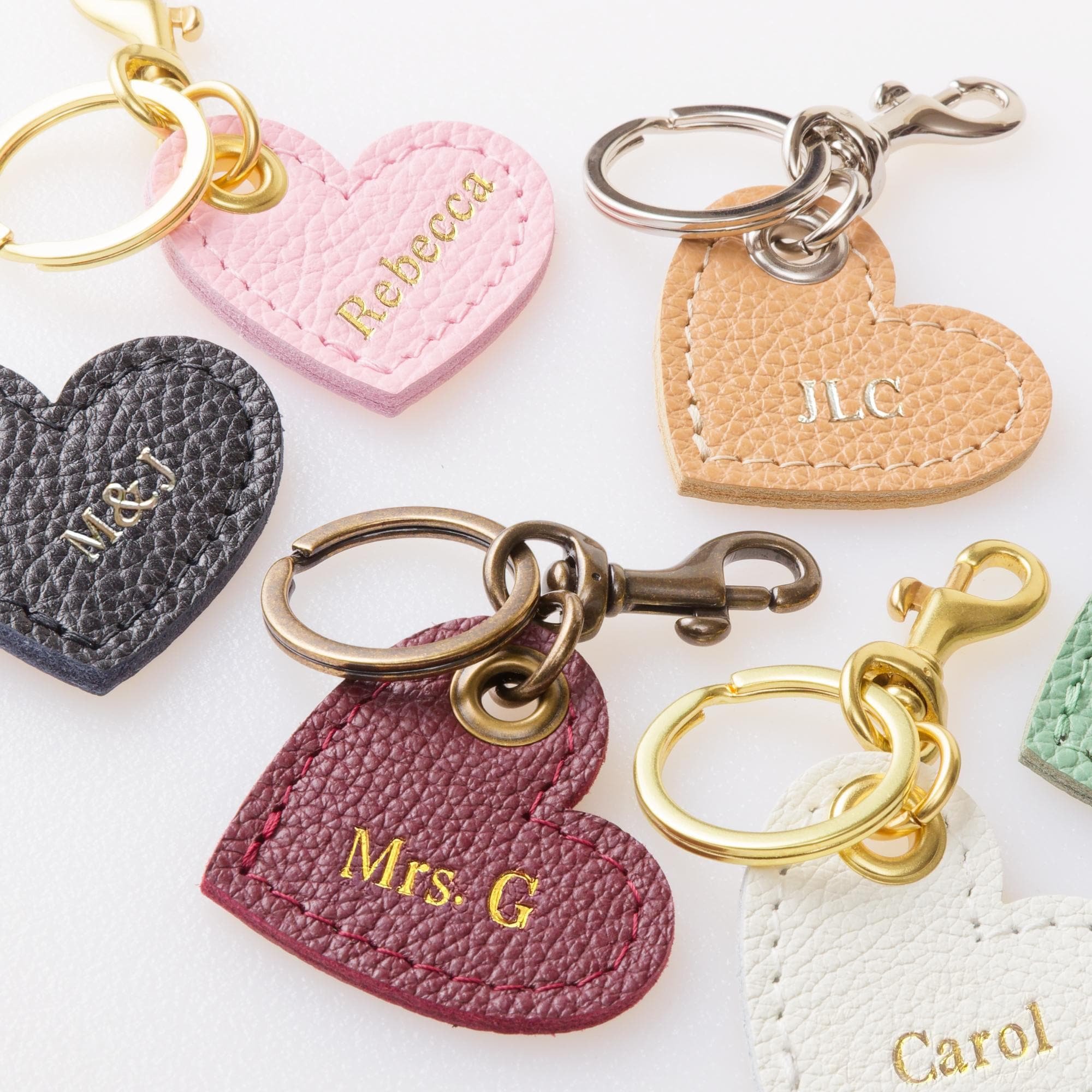 Variety of leather and hardware color heart shaped keychains on tabletop with personalization foil lettering.