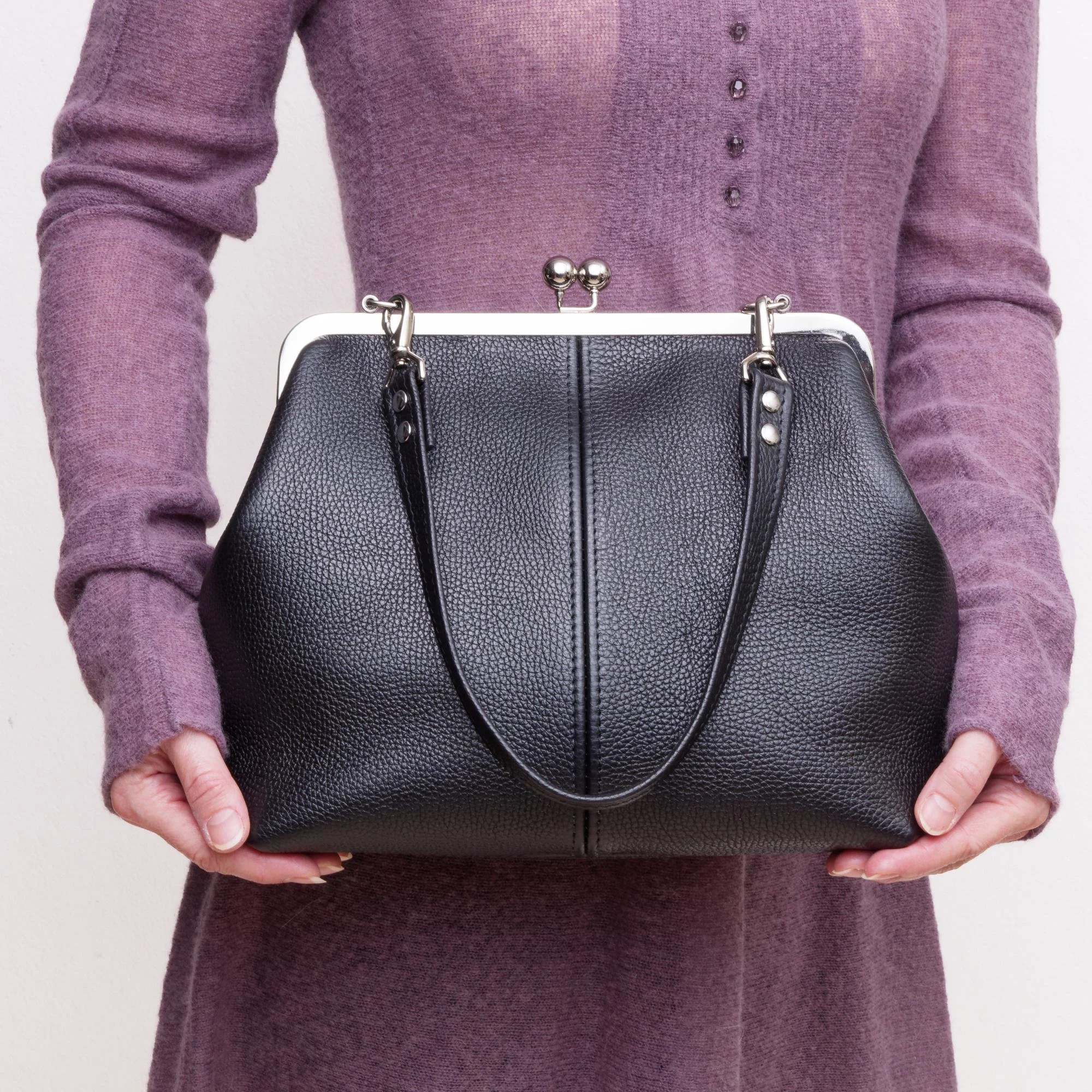 Model holding black pebble grain leather kiss lock handbag with silver finish hardware and shorty shoulder strap.