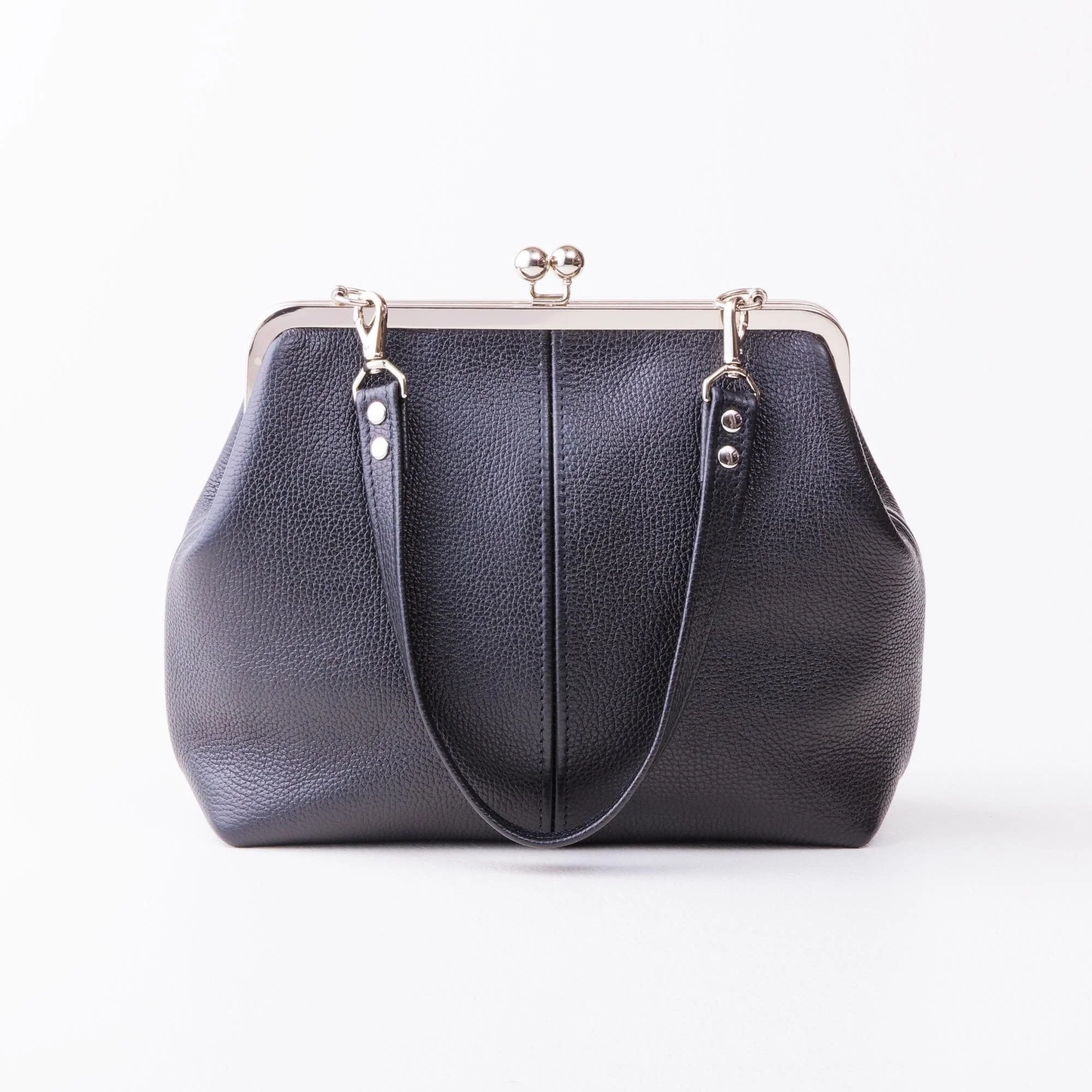 Front view of black pebble grain leather kiss lock handbag with silver finish hardware and shorty shoulder strap on tabletop.