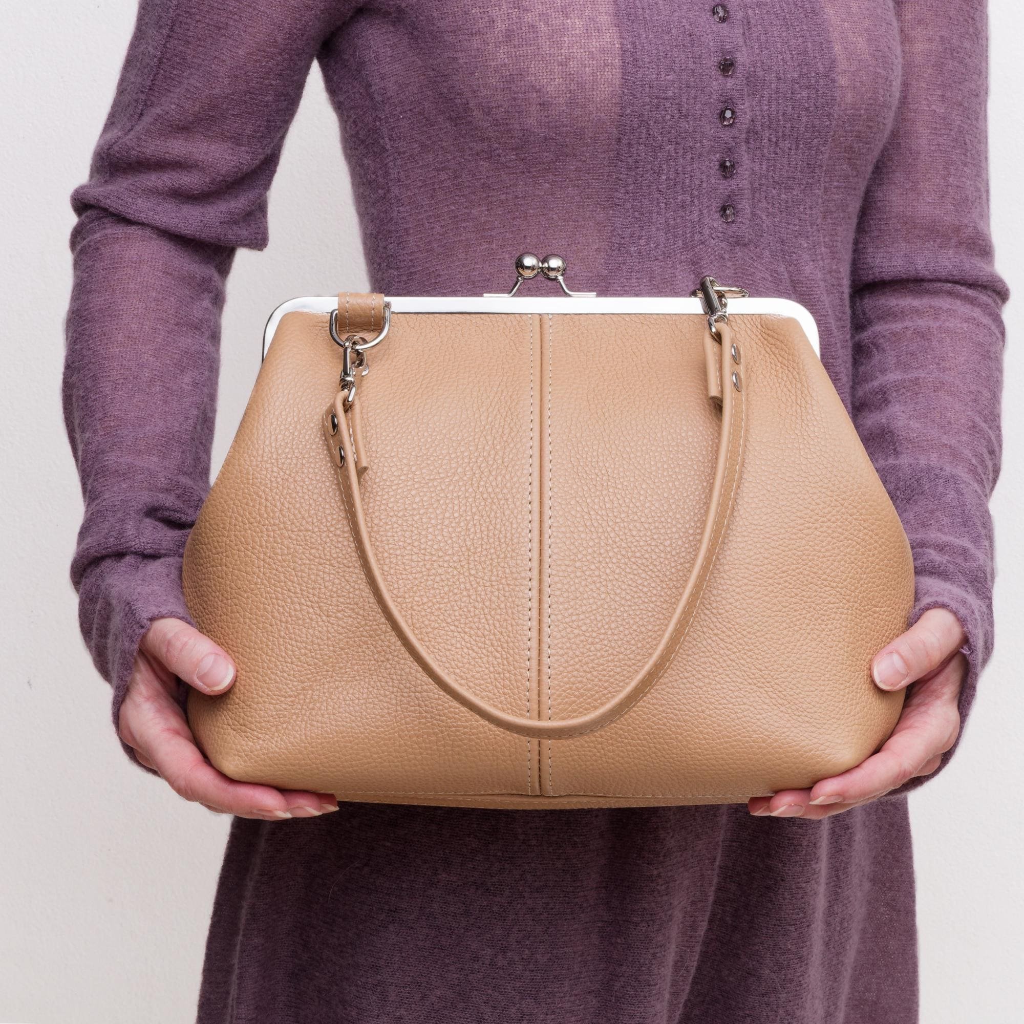 Model holding tan pebble grain leather kiss lock handbag with silver finish hardware and shorty shoulder strap.