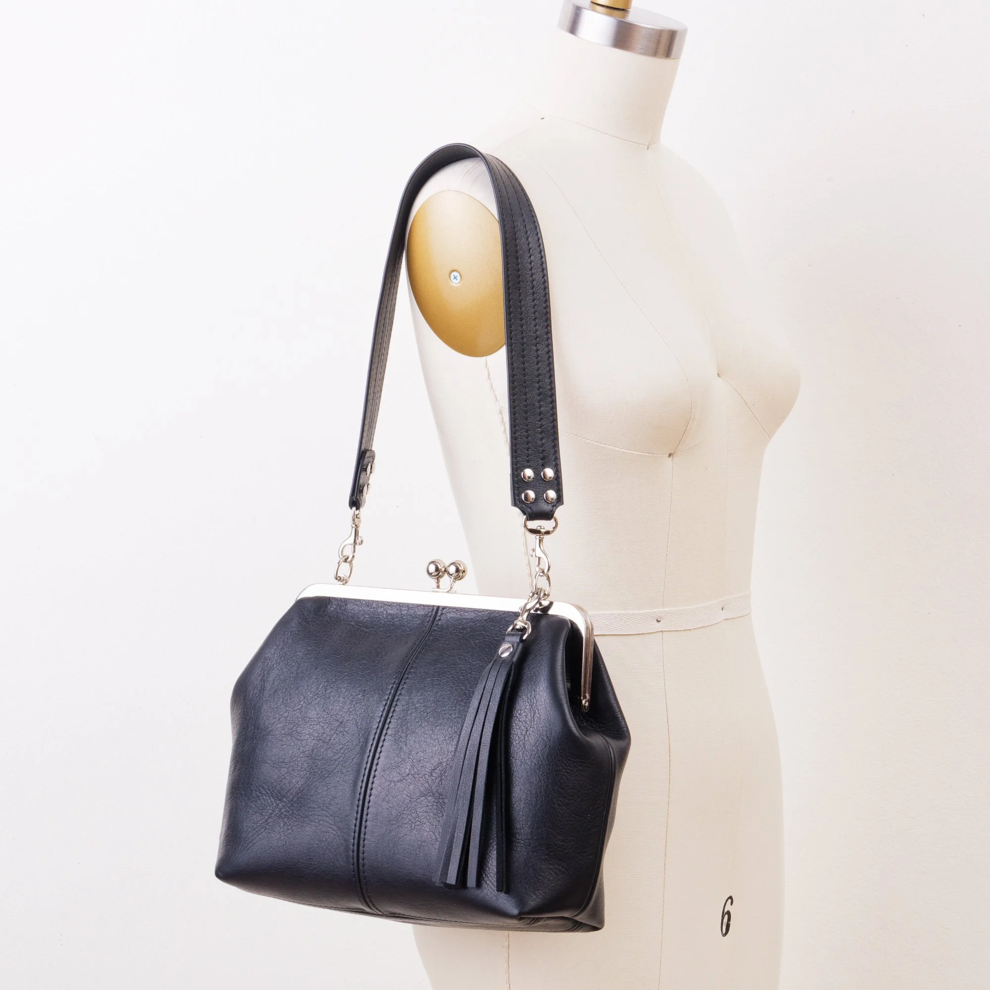 Black leather kiss lock handbag with silver finish hardware, 1.5 inch wide shoulder strap and tassel charm on mannequin.