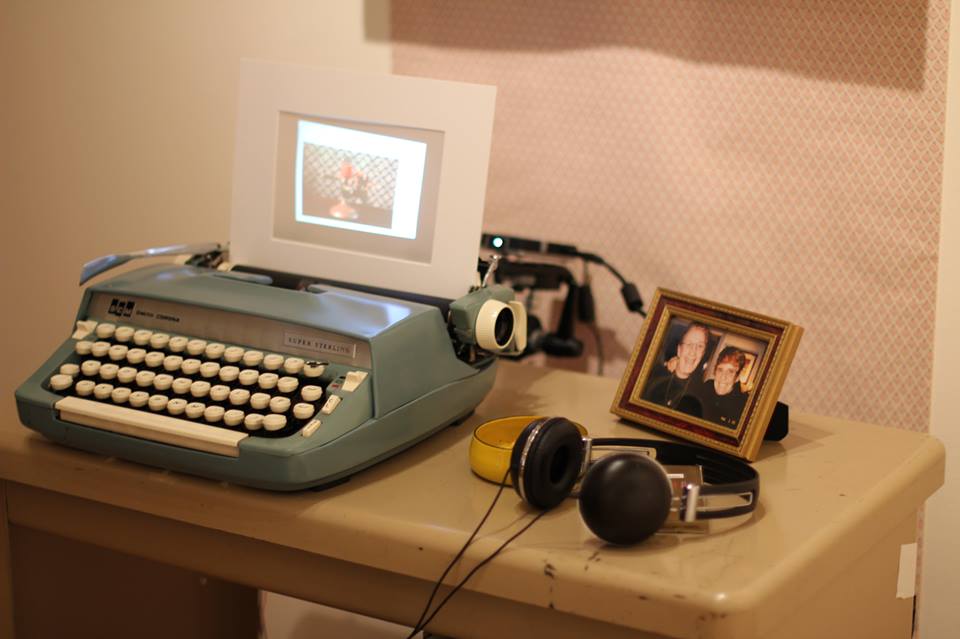 video is rear projected onto paper in a blue typewriter on a desk with headphones, ash tray, and framed picture of two older women