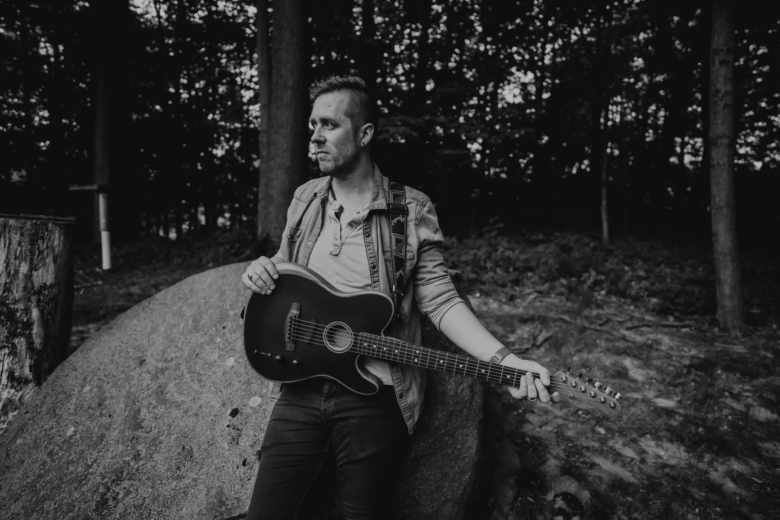A man with short hair standing outdoors in a wooded area, holding an acoustic guitar and looking to the left.