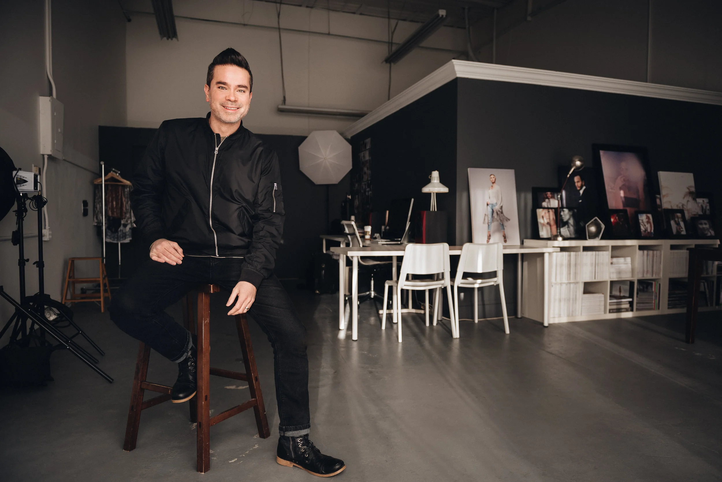 A man with dark hair and a short beard, wearing a black bomber jacket and jeans, sitting on a wooden stool in a photography studio. Behind him are photography equipment, a white table with various framed photographs, and studio lights. The studio has dark walls and a high ceiling.