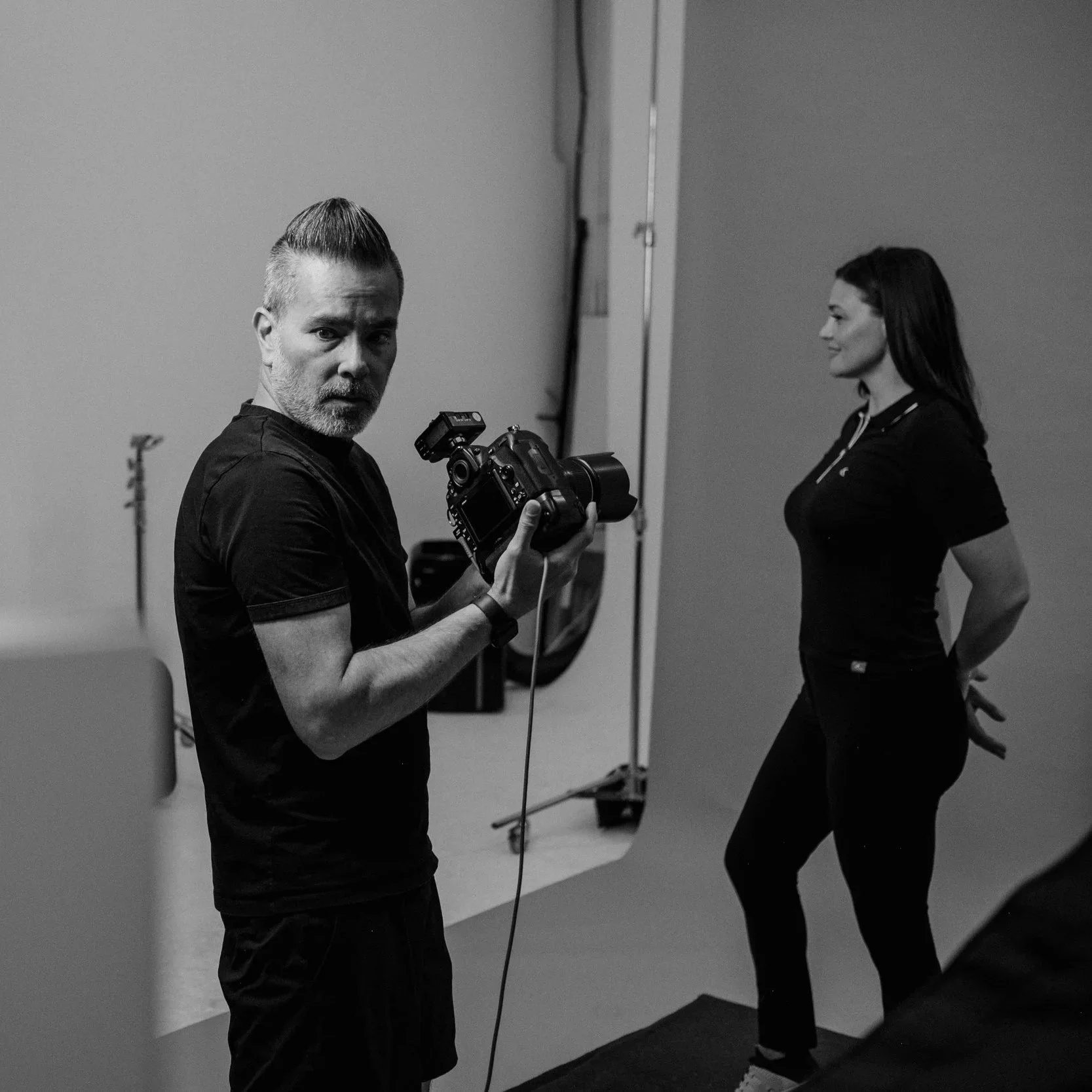 Black and white photo of a male photographer holding a camera, standing in front of a plain backdrop, with a woman in a black outfit and white sneakers posing.