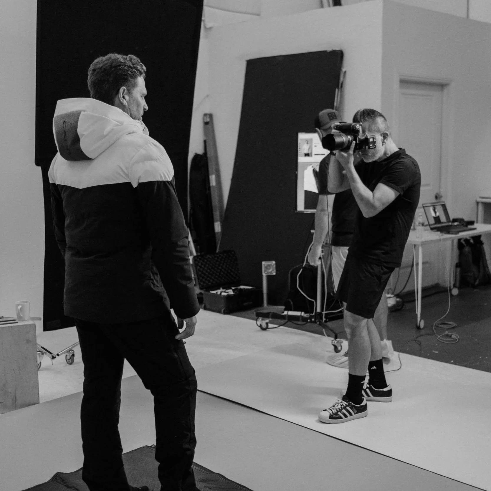 A photoshoot studio with two men, one holding a professional camera and the other in front of a backdrop. The man with the camera is wearing a black shirt, black shorts, and sneakers. The other man is dressed in a white and black jacket and black pan