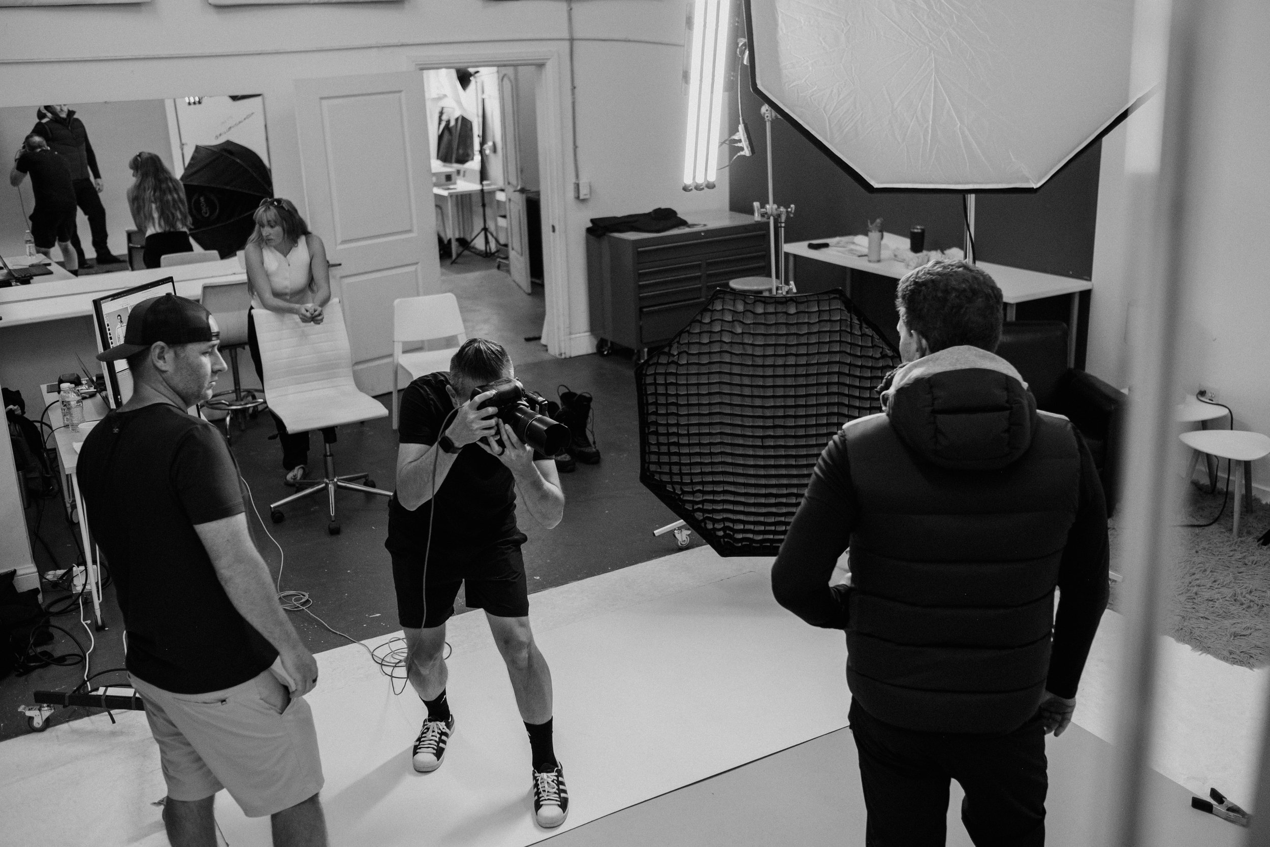 Black and white photo of a photography studio set, with a model walking on a white backdrop while photographers and crew members prepare and take pictures. Studio equipment such as lights, umbrellas, and a camera are visible.