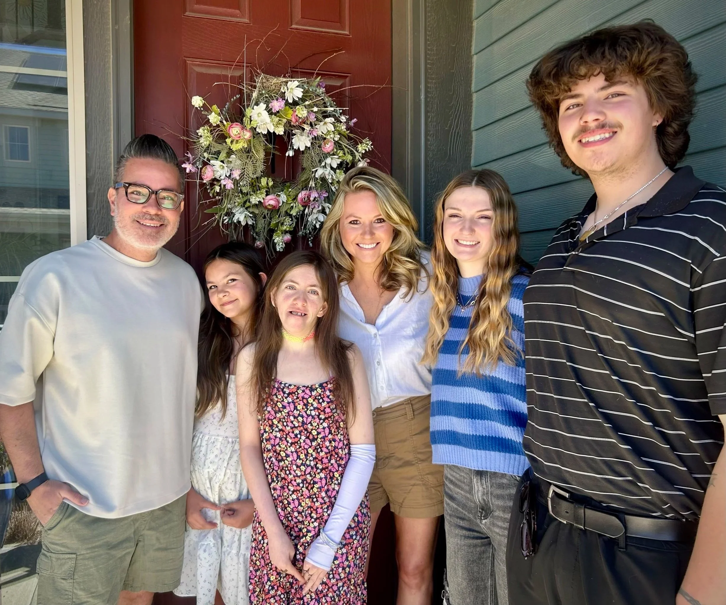 A group of seven people standing in front of a red door with a floral wreath, taking a selfie. They are smiling and appear happy.