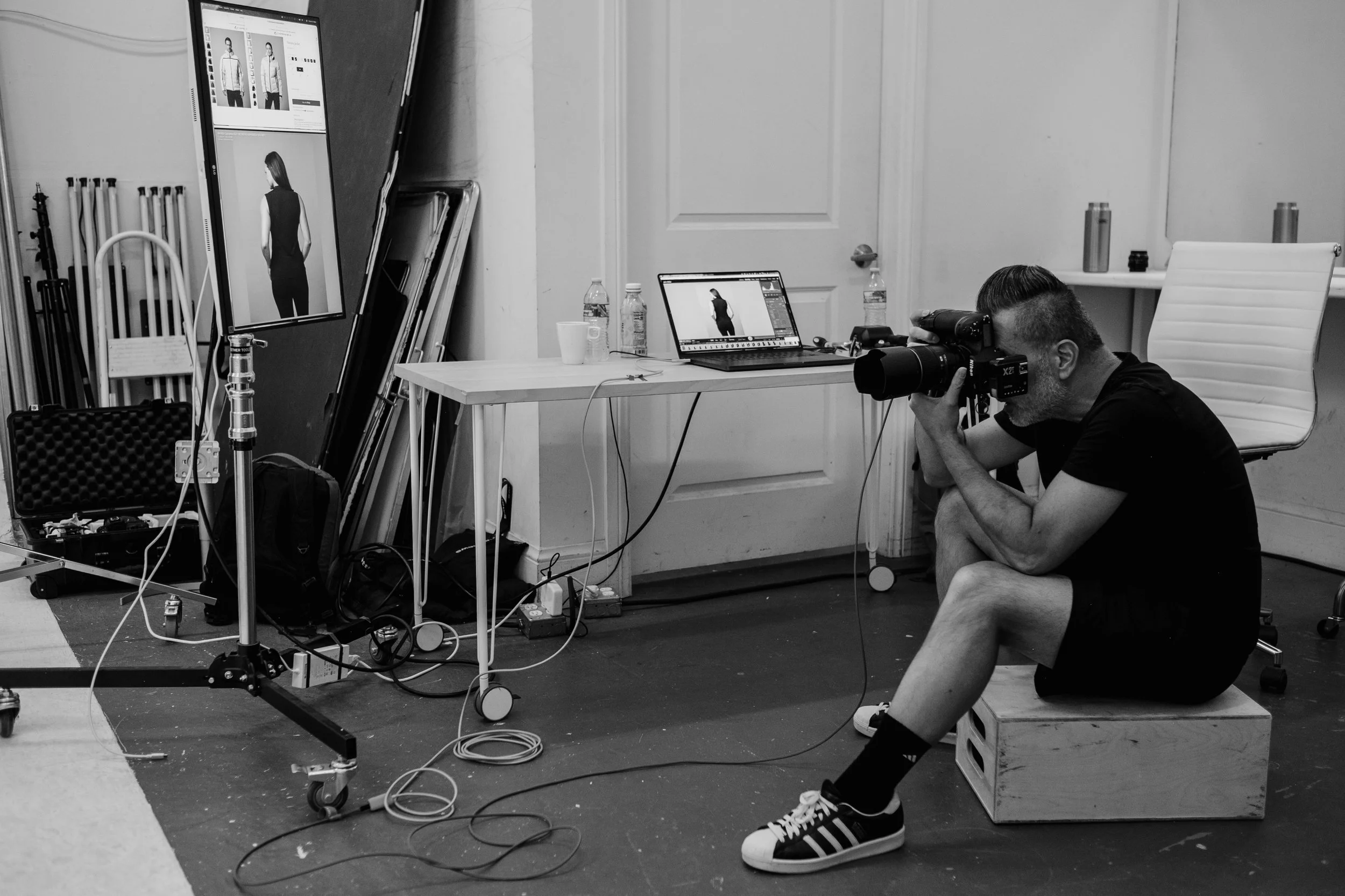 A photographer crouches on a wooden box with a camera to his eye, aiming at a model on a computer screen, in a photography studio filled with equipment and monitors displaying images of a woman in a black dress.