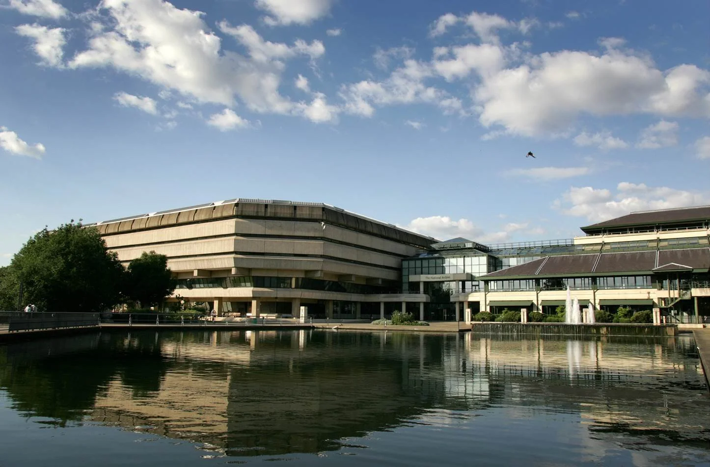 the national archives in kew, england on a sunny day with a reflecting pool in front and white clouds on a blue sky. Brutalist architecture heavier above than below, with a glass atrium joining two buildings.
