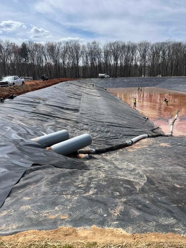 Black geomembrane liner across a large basin, with pipe penetrations on the slope and a crew working on the floor