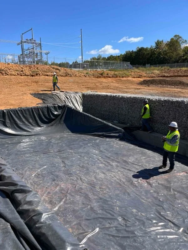 Black geomembrane liner laid in an excavation beside a gabion stone-basket wall, with crew on site