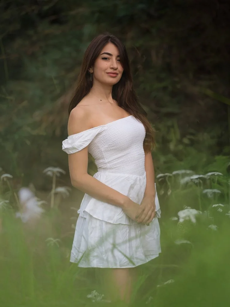 A young female model standing in green field and white flowers. She is wearing a white dress and looking at the camera while smiling..jpg