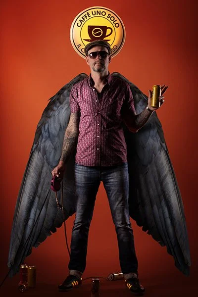 Photomontage einer Heiligenfigur. Ein Mann mit Taubenflügeln, Sonnenbrillen und Tätowierungen vor einem Leuchtschild eines Cafes. Er führt eine Bierdose an einer Hundeleine.