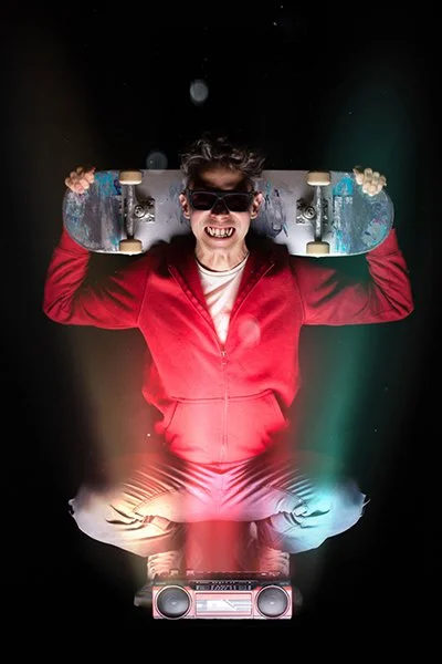 Photomontage einer Heiligenfigur.  Ein Mann mit Sonnenbrille und Reißzähnen kniet hinter einem Radio. Er hält ein Skateboard und grinst breit.