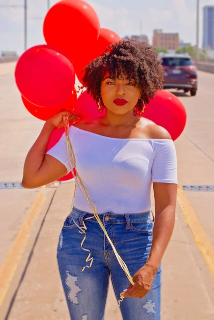 Woman holding a bunch of red balloons, wearing an off-shoulder white top and ripped jeans, standing outdoors on a parking lot with city buildings in the background.