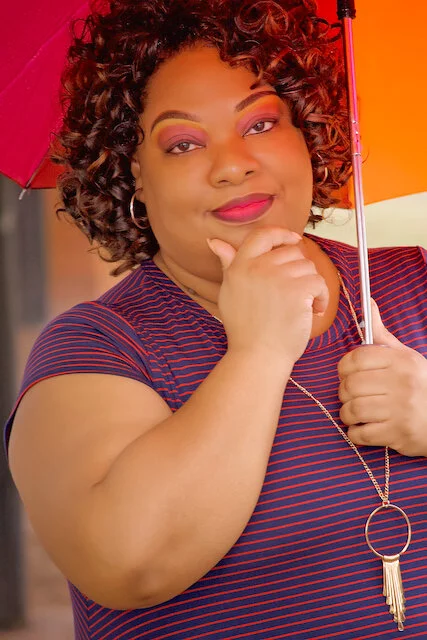 A woman with curly hair holding an orange umbrella, wearing a striped shirt and a necklace, posing with her hand on her chin.