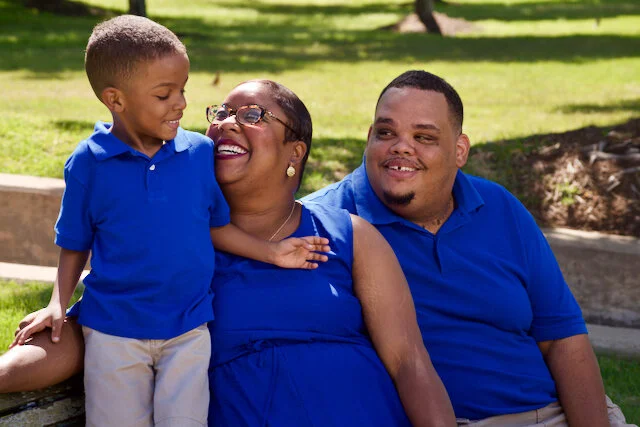 A family of three sitting on a park bench, smiling and enjoying a sunny day. The child is standing on the bench, leaning towards the woman in the middle, who is laughing. All are dressed in blue shirts.