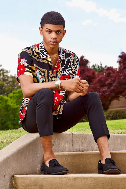 A young man sitting on outdoor steps in a park with trees and a blue sky in the background. He is wearing a colorful patterned shirt, black pants, and black shoes.