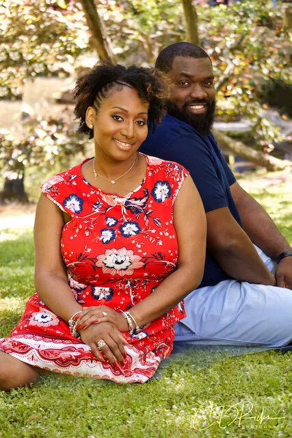 A smiling woman in a red floral dress sitting on grass next to a smiling man in a blue shirt and gray pants, outdoors with trees in the background.