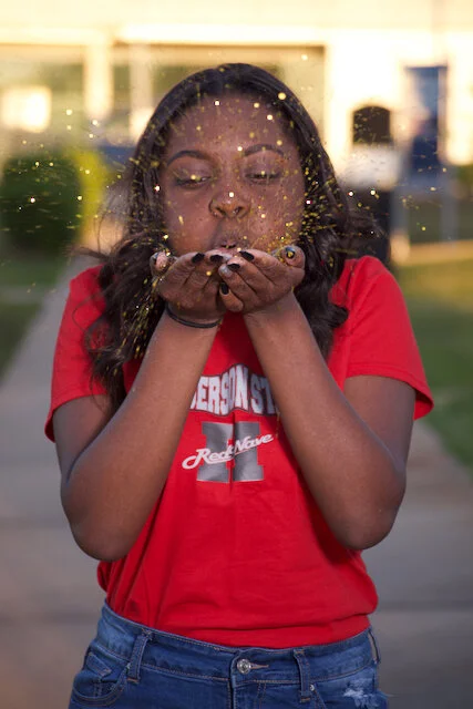 Young woman in a red t-shirt blowing glitter from her hands outdoors during sunset.