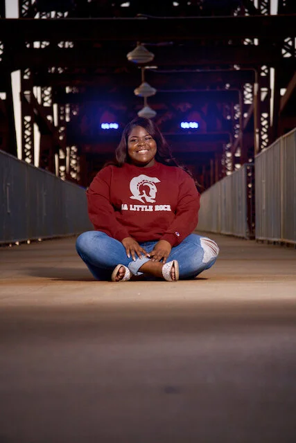 Young woman sitting cross-legged on a bridge, smiling at the camera, wearing a red sweatshirt with a helmet logo and the text "MA LITTLE ROCK." The bridge has industrial-style lighting and metal railings.