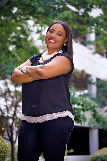 A woman standing outdoors with arms crossed, smiling, wearing a black top with lace trim, pearl necklace, and earrings, with greenery in the background.