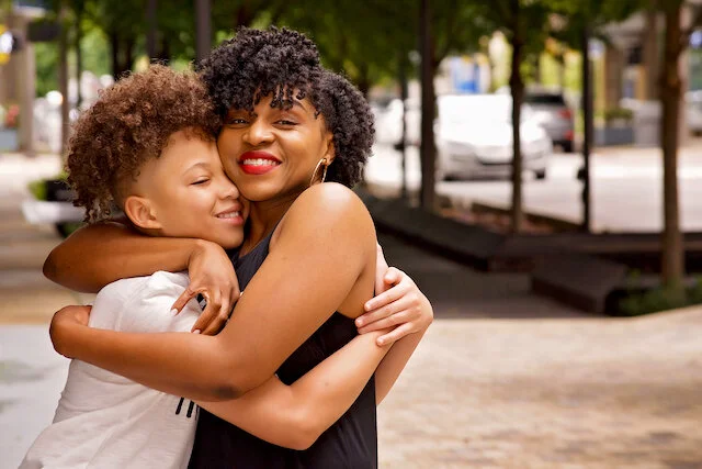 A woman and a young girl hugging and smiling outdoors in a park