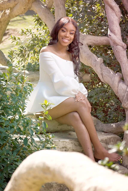 A young woman with long dark hair, sitting outdoors on a stone step among green bushes and trees, wearing an off-the-shoulder white dress, smiling at the camera.