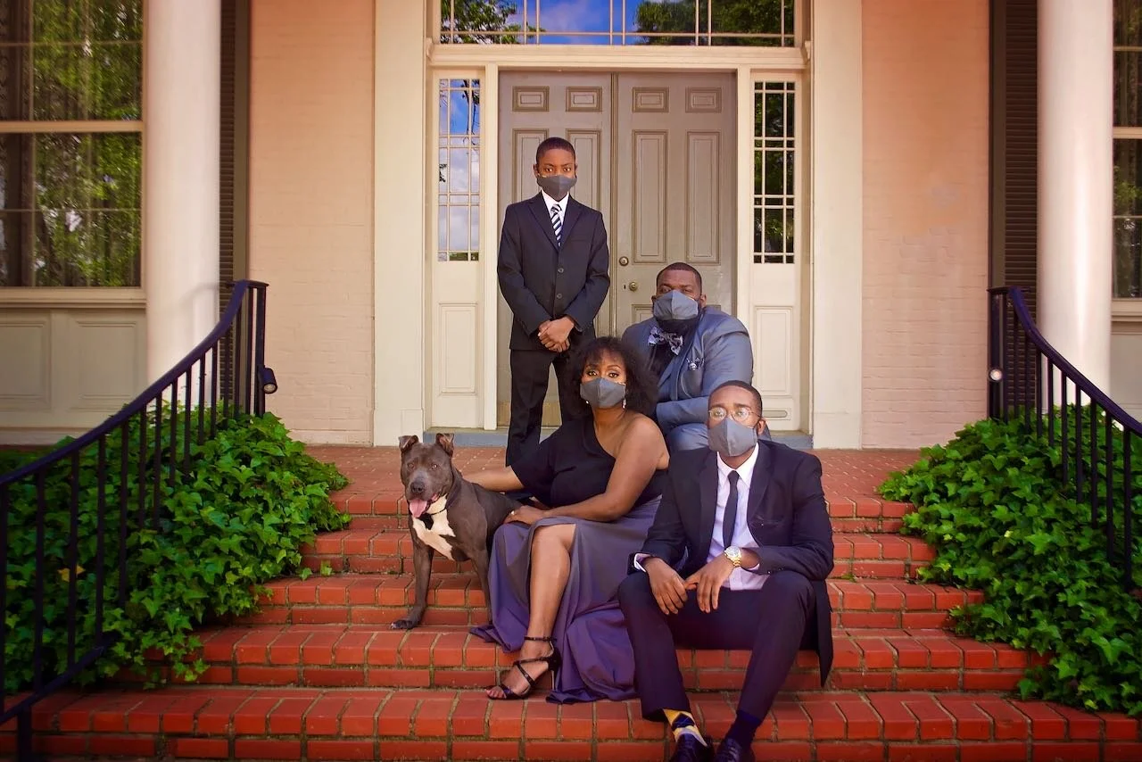 Family of four and a dog sitting on steps outside a house, all wearing masks. The family is dressed formally, with the woman in a black dress, the man in a black suit, the boy in a suit with a tie, and the man in a gray suit with a bow tie. The dog i