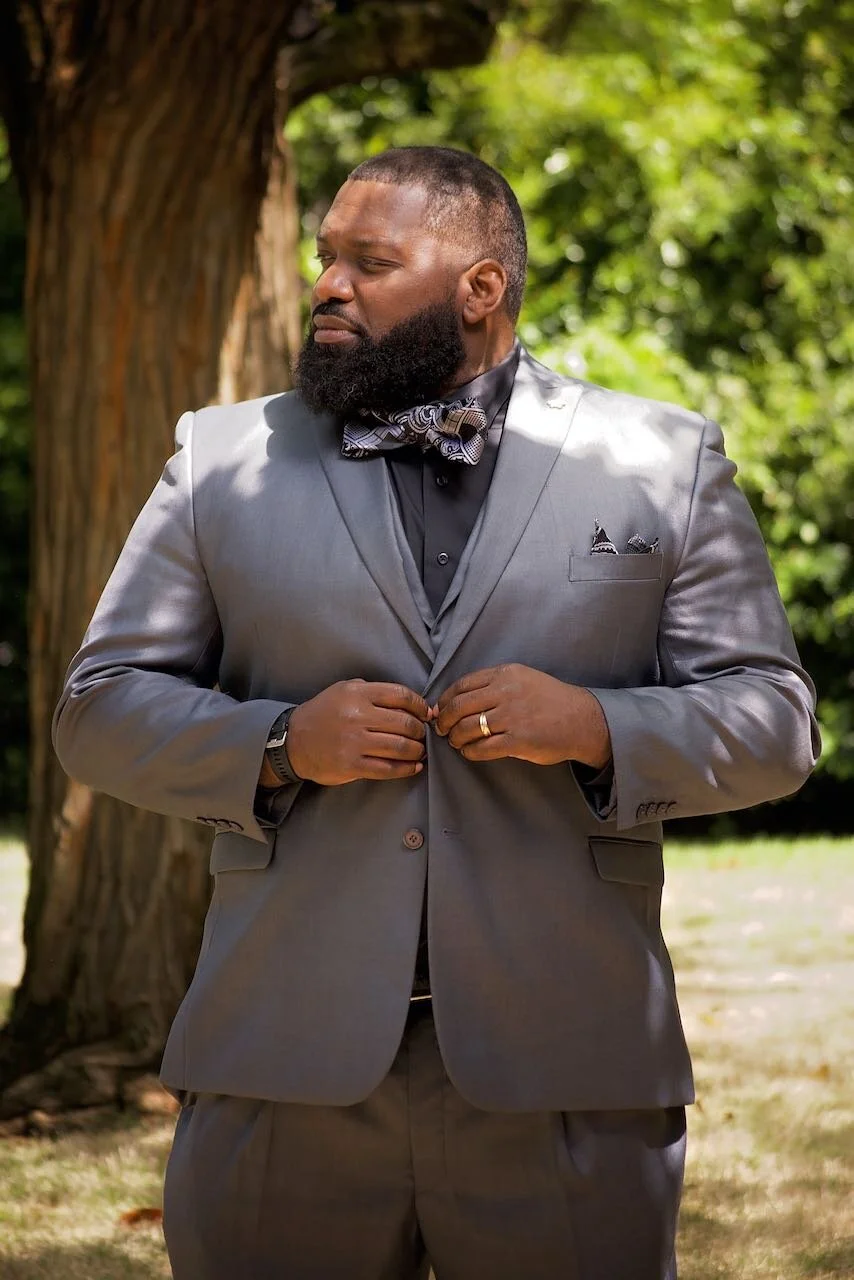 Man wearing a grey suit adjusting his jacket outdoors with green trees in the background.