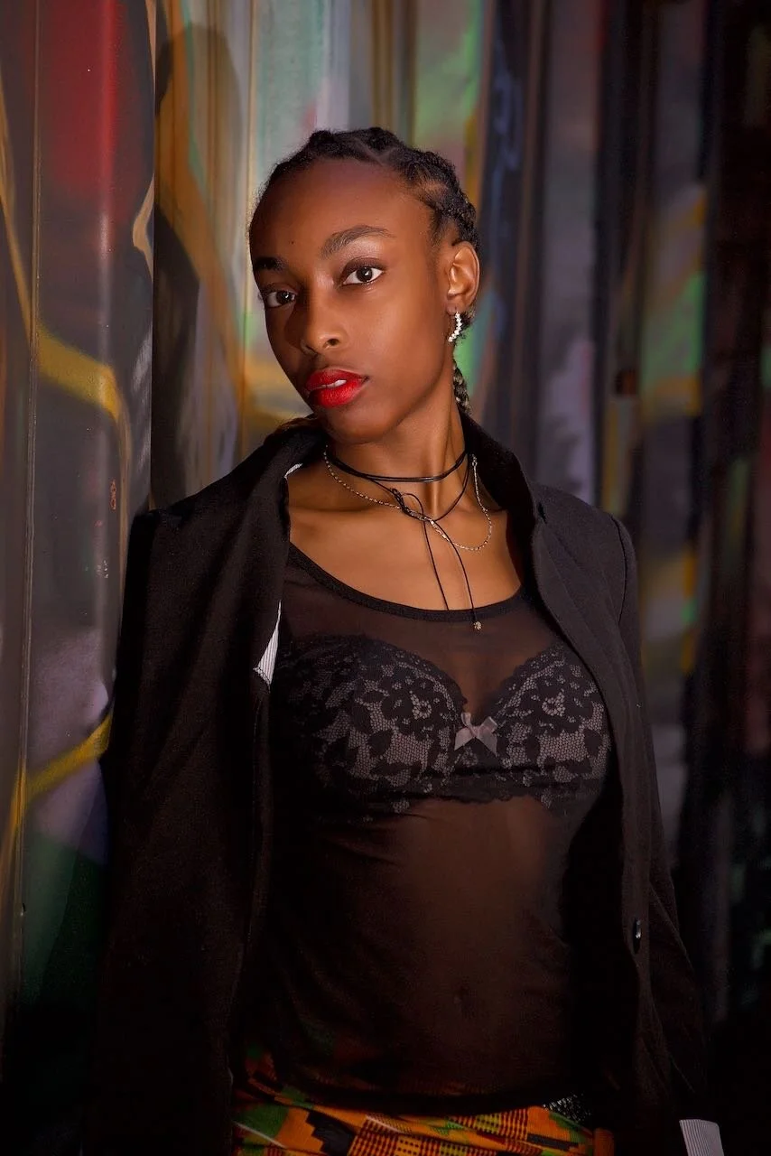 A young African American woman with braided hair, wearing a black sheer top with lace details, red lipstick, earrings, and layered necklaces, standing against a colorful graffiti wall.