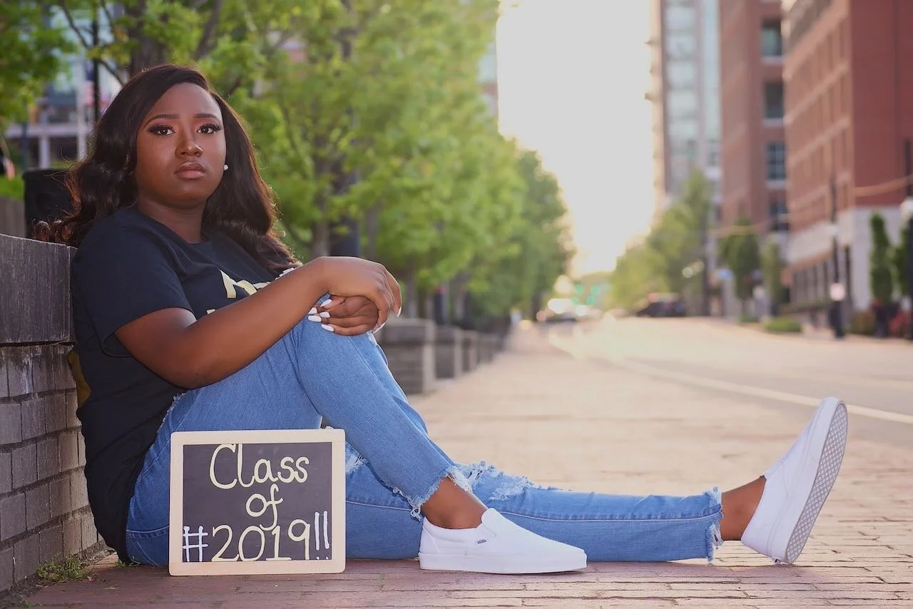 A young woman sitting on a sidewalk next to a chalkboard that says "Class of #2019!!" in an urban setting with trees and buildings in the background.
