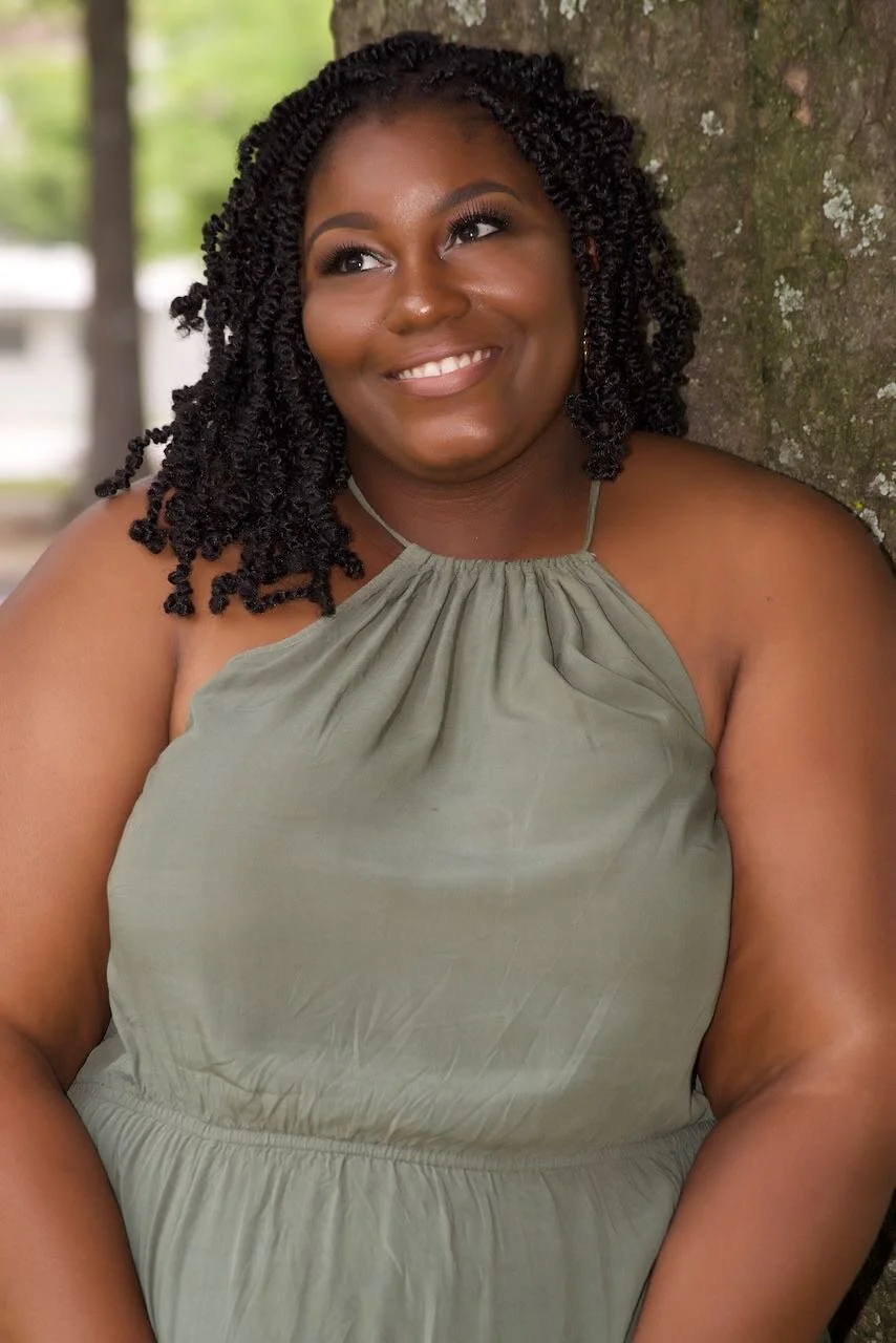A woman with dark curly hair smiling while leaning against a tree in an outdoor setting.