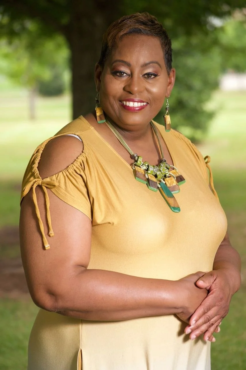 A smiling woman with short hair, wearing a yellow top with shoulder cut-outs and tied sleeves, accessorized with a colorful statement necklace and matching earrings, standing outdoors with a blurred green park background.