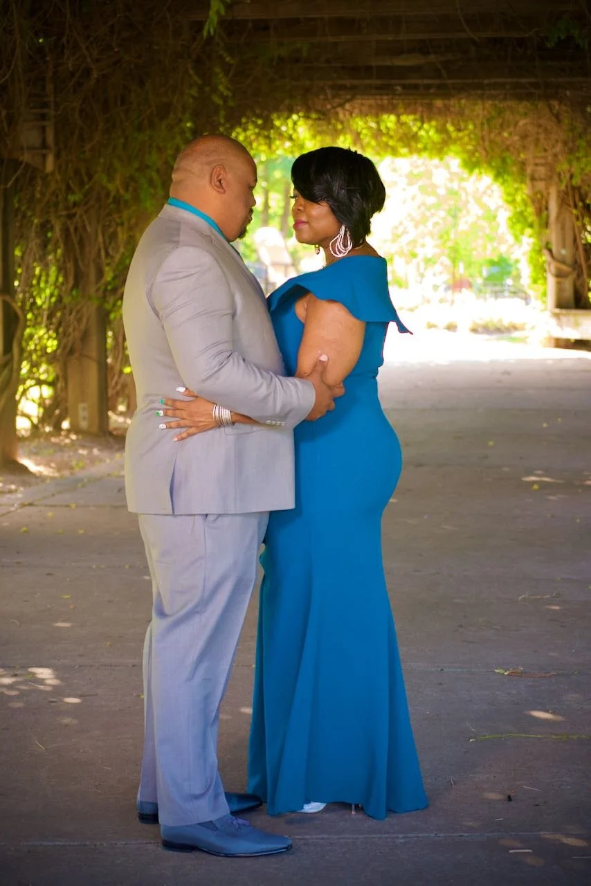 A couple with dark skin is standing close together with their foreheads touching. The man is wearing a light gray suit and the woman is wearing a long blue dress. They are outdoors under a tree-lined archway, with sunlight filtering through the leave