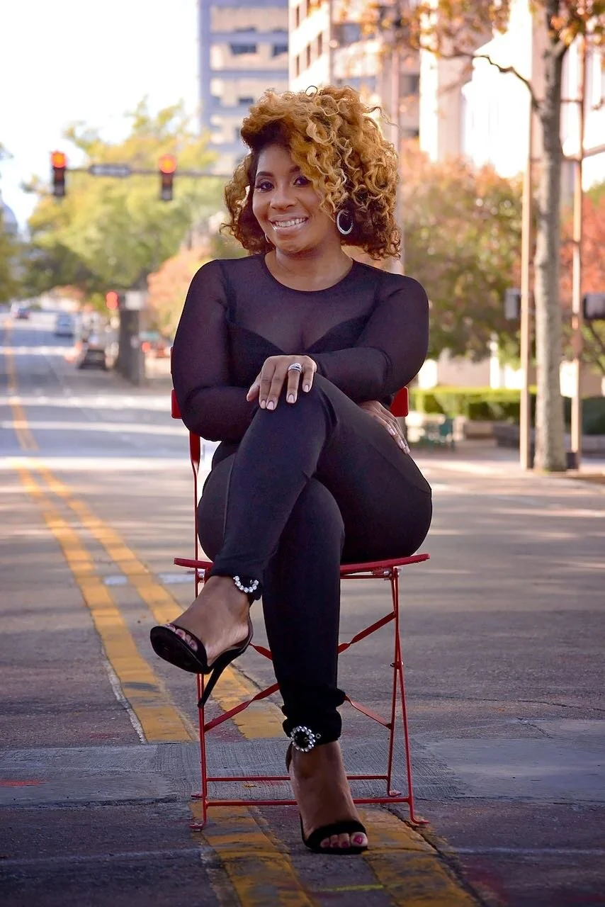 A woman with curly blonde hair sitting on a red chair in the middle of a city street, smiling and wearing black clothing and high heels.