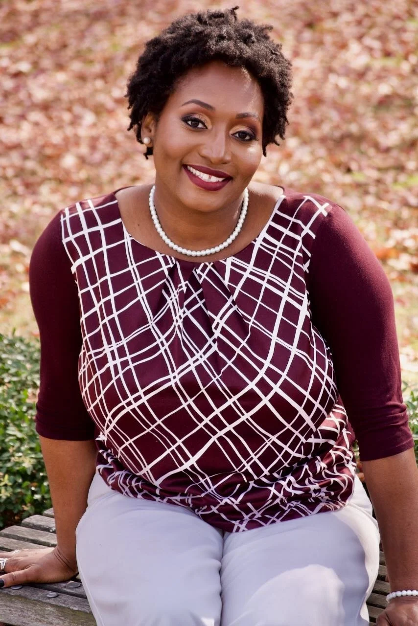 A woman smiling outdoors, wearing a burgundy and white patterned top, white pants, pearl necklace, and pearl earrings, sitting on a wooden bench.