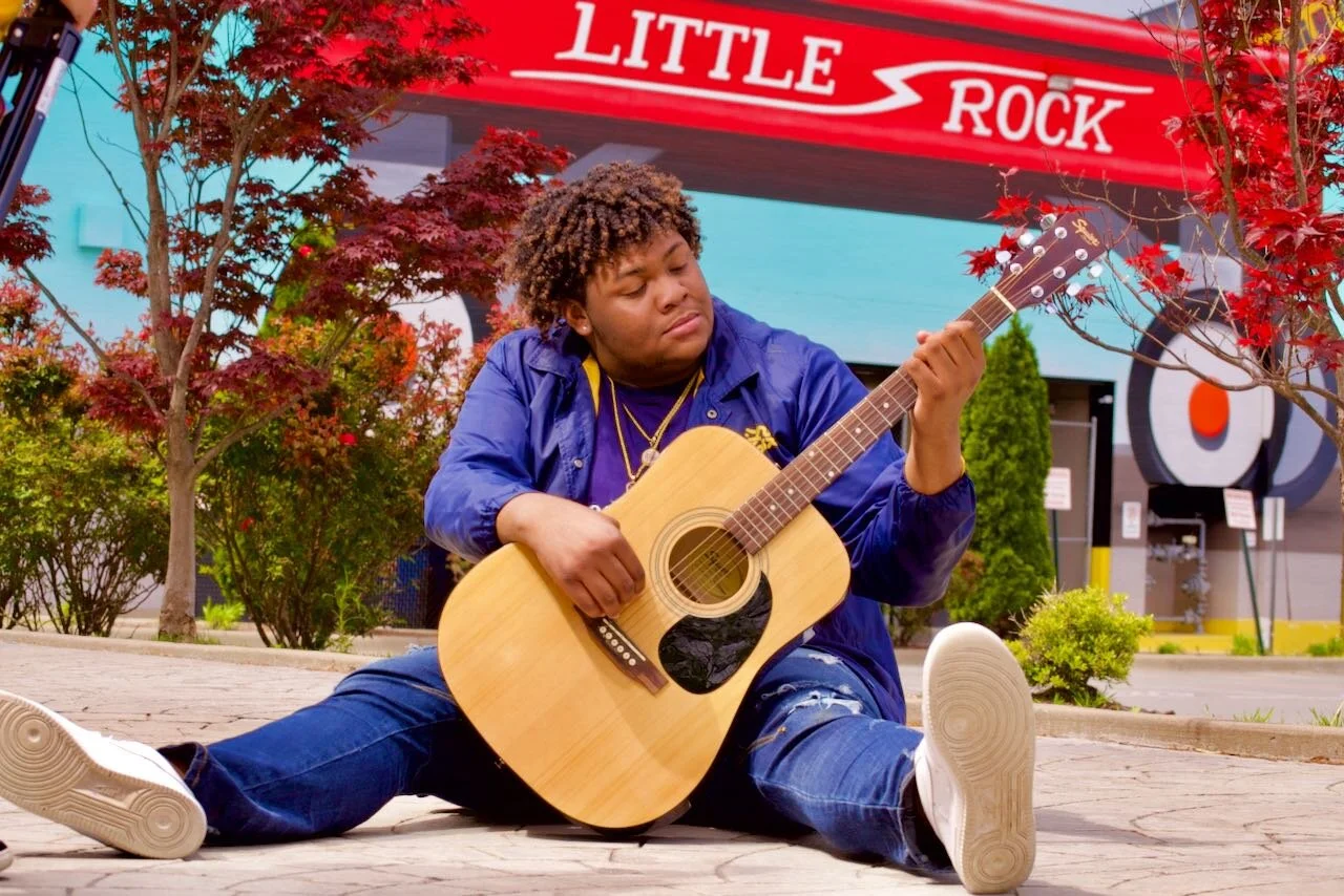 A young man sits on the sidewalk playing an acoustic guitar, surrounded by trees and bushes, with a Little Rock sign in the background.
