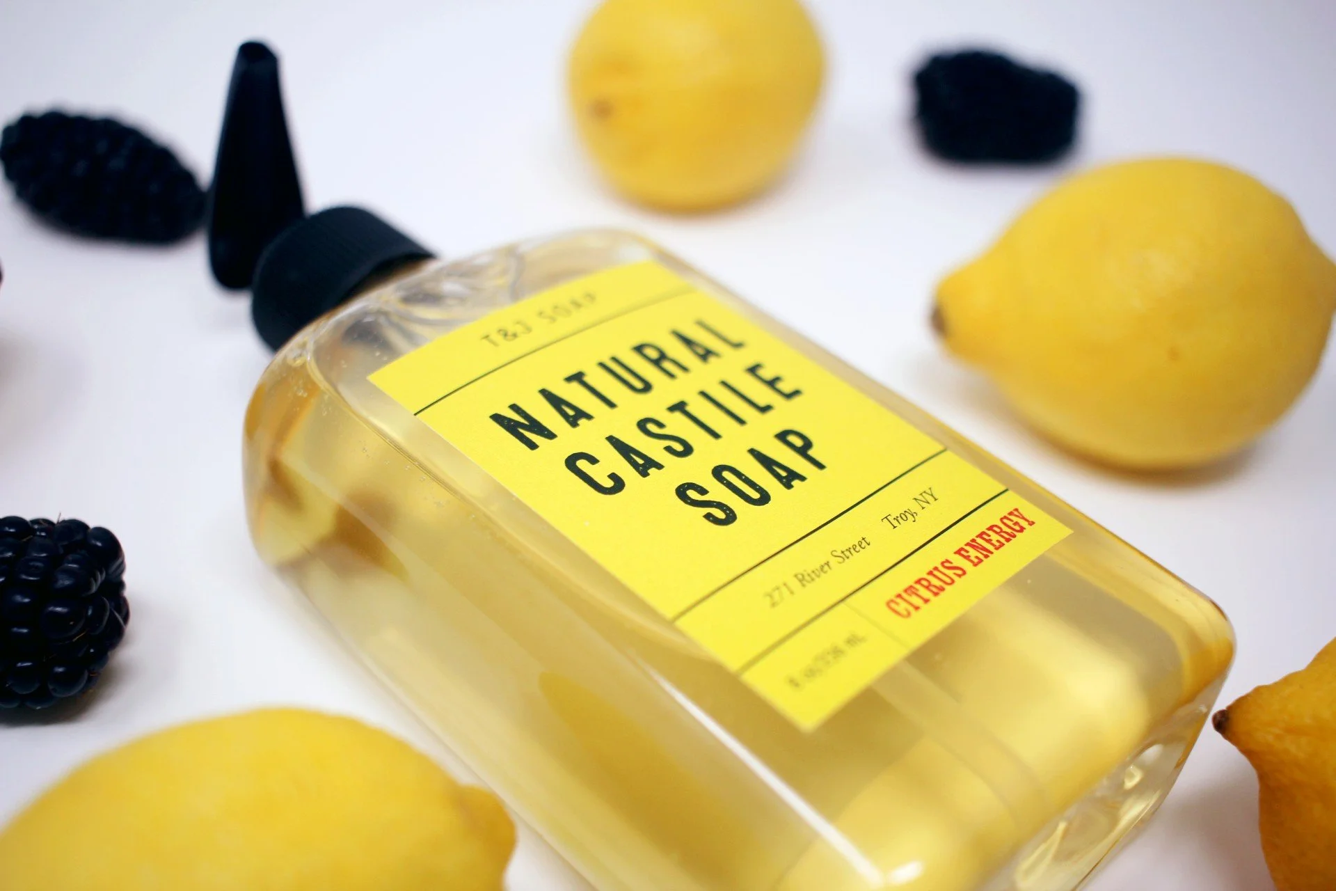 8 Ounce Bottle of Citrus Energy Castile Soap. Bottle has yellow label and is surrounded by blackberries and lemons. 