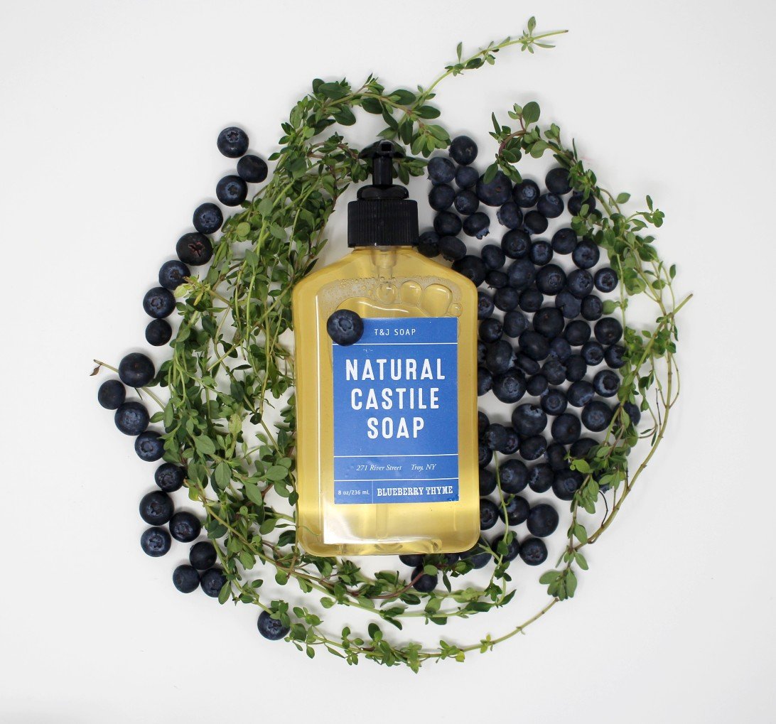 Bottle of Blueberry Thyme Castile Liquid Soap laying on top of a bed made of real blueberries and thyme.